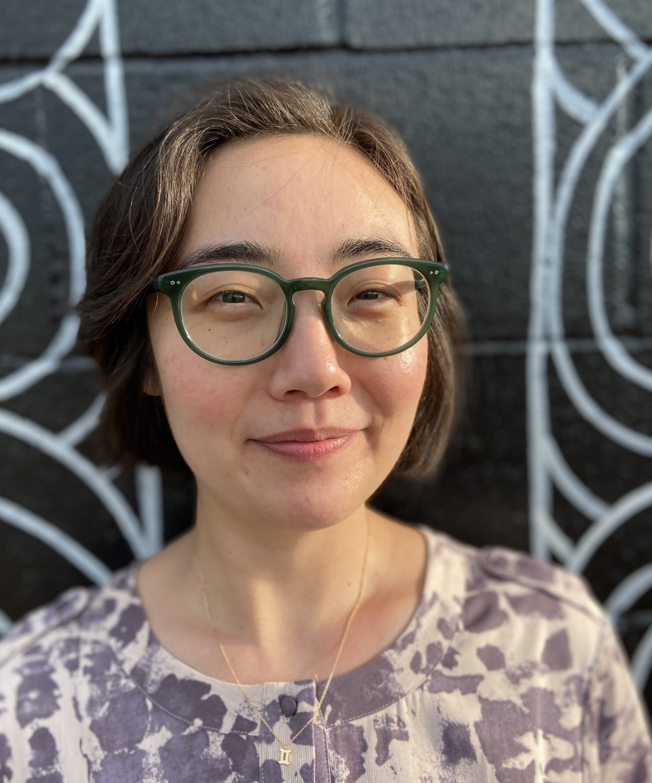 Korean American woman with short hair and green glasses stands against a black mural.